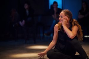 A long-haired White woman sits on the ground, demonstrating a contemporary dance move. / Une femme Blanche aux cheveux longs est assise et montre un pas de danse contemporain.