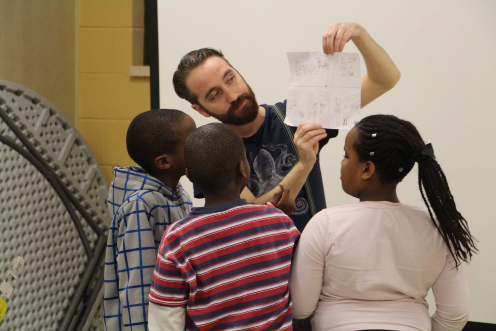 A White man observes a piece of paper with pencil drawings on it that he holds up so the three Black children in front of him can also see it. / Un homme Blanc observe une feuille avec des dessins au crayon qu’il tient en l’air pour que les trois enfants Noirs devant lui peuvent aussi la regarder.