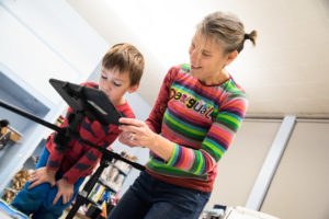 A White woman smiles as she shows a young boy something on an iPad attached to a tripod. / Une femme Blanche sourit en montrant quelque chose sur un iPad attaché à un trépied à un petit garçon.