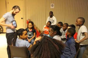 A White man holds an iPad in front of him, standing in front of a group of seated children. / Un homme Blanc présente un iPad à un groupe d’enfants assis devant lui.