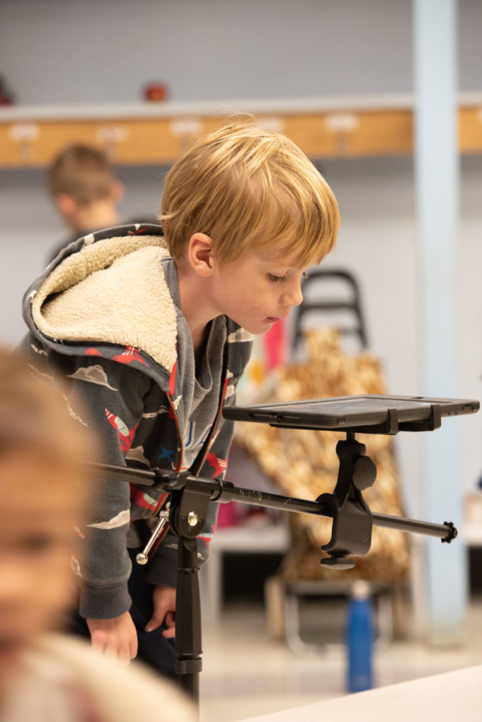 A young boy leans down to look at an iPad attached to a tripod, facing the ground. / Un petit garçon se penche au dessus d’un iPad attaché à un trépied, face au sol.