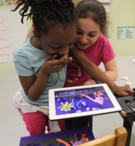 Two young girls smile over an iPod perched above a collage as they take pictures of it. / Deux petites filles sourient en voyant le collage qu’elles photographient avec un iPad perché au dessus.