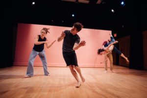 Two dancers sway towards the left in the foreground while a third dancer holds a fourth as they do the splits in midair. / Deux danseurs oscillent vers la gauche alors qu’un troisième tient un quatrième qui fait le grand écart dans les airs.