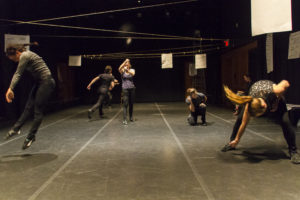 Five contemporary dancers move in different ways on a set. / Cinq danseurs contemporains dansent parmi un décor.