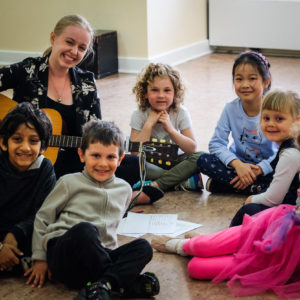 A White woman holding a guitar sits on the floor with six young children. / Une femme Blanche, une guitare a la main, est assise par terre avec six jeunes enfants.