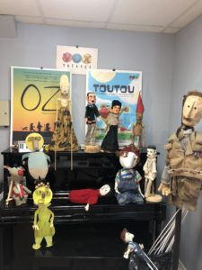 A black piano with puppets on top and posters behind it. / Un piano noir avec des marionnettes et des affiches derrière.