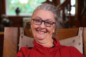 A blue eyed White woman with salt and pepper hair smiles at the camera. / Une femme Blanche aux yeux bleus et cheveux noirs et blancs sourit.