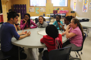 A man talks to girls around a table in a classroom. / Un homme parle à des filles autour d’une table dans une classe.