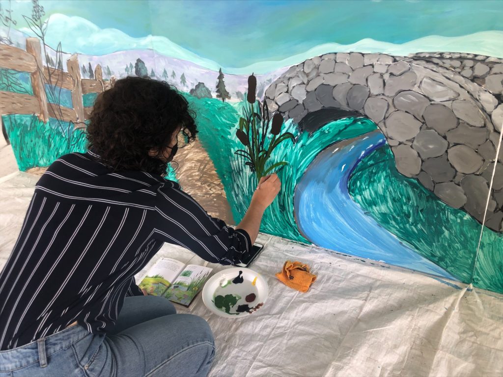 Nicole Belanger in a black and white striped blouse, light blue jeans and dark brown curly hair. There is a book and plate of paint colours in front of her as she faces the wall painting cattails.