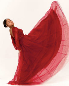 A young Black girl in a long red dress with tulle and polka dots kicks her foot straight into the air in front of her with her toes pointed, her back tilted backwards, head turned towards the camera, and holding her hands behind her back. / Une jeune fille Noire dans une longue robe rouge en tulle avec des petits pois lève sa jambe en l’air devant elle, le pied pointé, le dos courbé en arrière, la tête tournée vers l’objectif et tenant ses mains dans son dos.