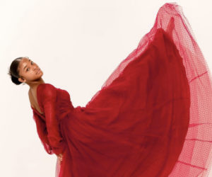 A young Black girl in a long red dress with tulle and polka dots kicks her foot straight into the air in front of her with her toes pointed, her back tilted backwards, head turned towards the camera, and holding her hands behind her back. / Une jeune fille Noire dans une longue robe rouge en tulle avec des petits pois lève sa jambe en l’air devant elle, le pied pointé, le dos courbé en arrière, la tête tournée vers l’objectif et tenant ses mains dans son dos.