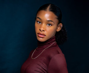 A young Black girl in a burgundy turtleneck and gold chain, with her hair slicked back into a ponytail, turns her head to face the camera, looking straight into the lens. / Une jeune fille Noire porte un col roulé bordeaux et une chaîne dorée, ses cheveux en queue-de-cheval, et tourne la tête vers la caméra en regardant droit dans l’objectif.