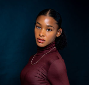 A young Black girl in a burgundy turtleneck and gold chain, with her hair slicked back into a ponytail, turns her head to face the camera, looking straight into the lens. / Une jeune fille Noire porte un col roulé bordeaux et une chaîne dorée, ses cheveux en queue-de-cheval, et tourne la tête vers la caméra en regardant droit dans l’objectif.