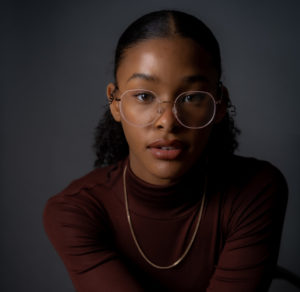 A young Black girl in rounded glasses, a burgundy turtleneck and gold chain, with her hair slicked back into a ponytail, faces the camera, looking straight into the lens. / Une jeune fille Noire porte des lunettes circulaires, un col roulé bordeaux et une chaîne dorée, a une queue-de-cheval, et fait face à la caméra, regardant droit dans l’objectif.