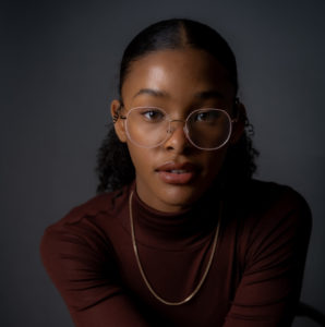 A young Black girl in rounded glasses, a burgundy turtleneck and gold chain, with her hair slicked back into a ponytail, faces the camera, looking straight into the lens. / Une jeune fille Noire porte des lunettes circulaires, un col roulé bordeaux et une chaîne dorée, a une queue-de-cheval, et fait face à la caméra, regardant droit dans l’objectif.