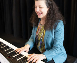 A dark haired, brown-eyed White woman smiles while playing the keyboard. / Une femme Blanche aux yeux et cheveux bruns sourit en jouant du piano électrique.