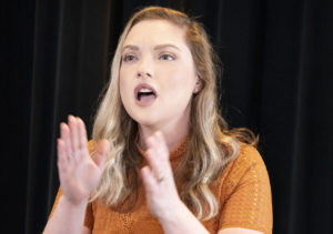 A blue-eyed, blonde haired White woman sings, holding her hands out in front of her. / Une femme Blanche aux yeux bleus et cheveux blonds chante, ses mains devant elle.