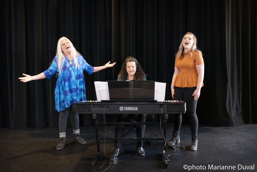 A White woman plays the keyboard, flanked on either side by two singing White women. / Une femme Blanche joue le piano électrique, deux femmes Blanches chantant à ses côtés.