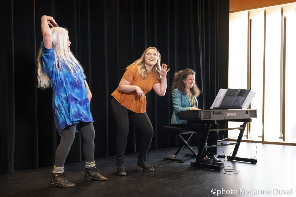 Two White women act things out as a third plays the keyboard. / Deux femmes Blanches font du théâtre alors qu’une troisième joue du piano électrique.