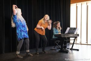 Two White women act things out as a third plays the keyboard. / Deux femmes Blanches font du théâtre alors qu’une troisième joue du piano électrique.