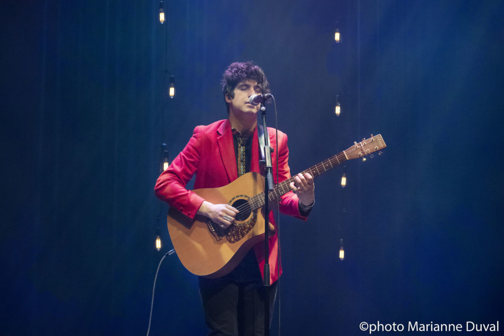 A black-haired man in a red blazer plays guitar and sings into a microphone. / Un homme aux cheveux noirs, portant un blazer rouge, joue de la guitare et chante dans un microphone.