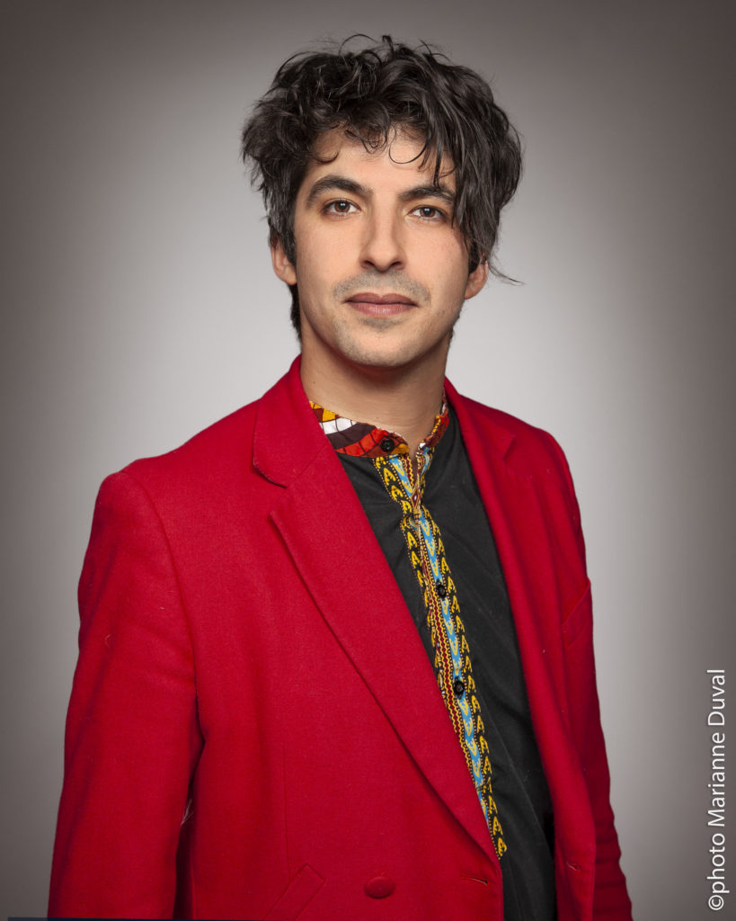 A black-haired man in a red blazer looks at the camera. / Un homme aux cheveux noirs, portant un blazer rouge, regarde la caméra.