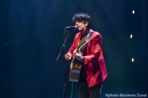 Seen from the side, black-haired man in a red blazer plays guitar and sings into a microphone. / Vu de profil, un homme aux cheveux noirs, portant un blazer rouge, joue de la guitare et chante dans un microphone.