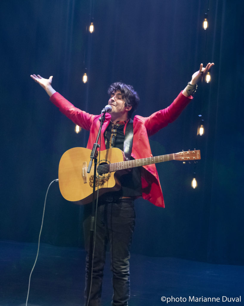 A black-haired man in a red blazer, his acoustic guitar strapped in front of him, smiles, raising his arms above his head, eyes closed. / Un homme aux cheveux noirs, portant un blazer rouge, sa guitare acoustique pendant de son épaule, sourit et lève les bras, les yeux fermés.