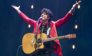 A black-haired man in a red blazer, his acoustic guitar strapped in front of him, smiles, raising his arms above his head, eyes closed. / Un homme aux cheveux noirs, portant un blazer rouge, sa guitare acoustique pendant de son épaule, sourit et lève les bras, les yeux fermés.