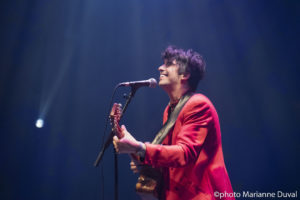 Seen from the side, a black-haired man in a red blazer plays guitar, smiling. / Vu de profil, un homme aux cheveux noirs, portant un blazer rouge, joue de la guitare en souriant.