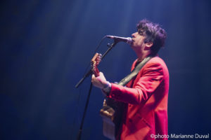 Seen from the side, a black-haired man in a red blazer plays guitar and sings into a microphone. / Vu de profil, un homme aux cheveux noirs, portant un blazer rouge, joue de la guitare et chante dans un microphone.