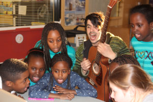 A man plays guitar and, with children, sings, their mouths open wide. / Un homme joue de la guitare et, avec des enfants, chante, leur bouches grandes ouvertes.