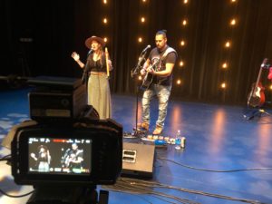 An Indigenous duo is filmed performing music on stage: the woman sings into a microphone while holding a flute, the man plays guitar. / Un duo Autochtone est filmé jouer de la musique sur une scène: la femme chante dans un micro et tient une flûte, l’homme joue de la guitare.