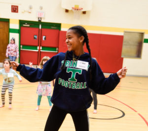 A young, smiling Black girl dances in a gymnasium as three young children follow along behind her. / Une jeune fille Noire souriante danse dans un gymnase alors que trois jeunes enfants reproduisent ses mouvements derrière elle.