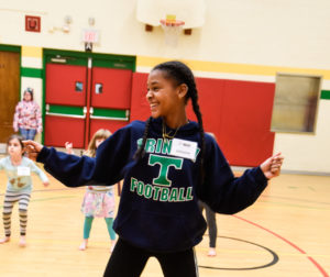 A young, smiling Black girl dances in a gymnasium as three young children follow along behind her. / Une jeune fille Noire souriante danse dans un gymnase alors que trois jeunes enfants reproduisent ses mouvements derrière elle.