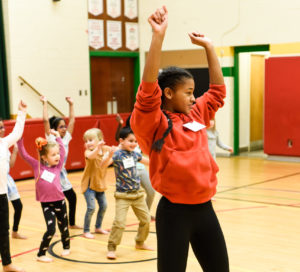 A young, smiling Black girl dances in a gymnasium with her back to a group of young children following along behind her. / Une jeune fille Noire souriante danse dans un gymnase, son dos à un groupe de jeunes enfants qui reproduisent ses mouvements.