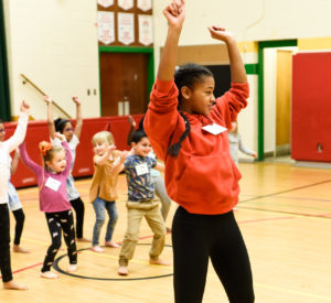 A young, smiling Black girl dances in a gymnasium with her back to a group of young children following along behind her. / Une jeune fille Noire souriante danse dans un gymnase, son dos à un groupe de jeunes enfants qui reproduisent ses mouvements.