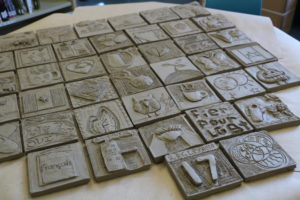 Rows of decorated unfired clay tiles lay on a table. / Des tuiles en argiles décorées non cuites sont en rangées sur une table.