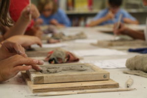 In the foreground, a child carves an unfired clay tile as others do the same in the background. / Un enfant grave une tuile en argile non cuite au premier plan pendant que d’autres font la même chose en arrière-plan.