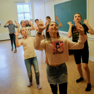 A few spaced out rows of children put their hands in the air as they look up to the ceiling. / Quelques rangées écartées d’enfants lèvent leurs mains en l’air en regardant vers le plafond.