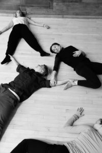 In black and white, four dancers lay on the floor, three holding onto each other. / En noir et blanc, quatre danseurs sont couchés par terre, trois d’entre eux se tiennent.