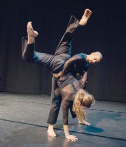 A dancer leans forward, holding an upside down danser, both all in black. / Un danseur se penche en avant en tenant un danseur à l’envers, les deux habillés en noir.