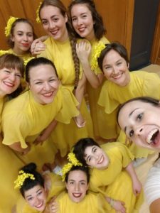 Nine White women and girls in yellow dresses smile for a selfie with a woman in a white shirt. / Neuf femmes et jeunes filles Blanches en robes jaunes sourient pour un selfie avec une femme en chandail blanc.