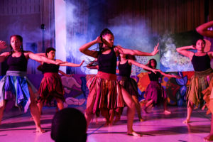 Several young women with dark skin wearing black tank tops and colourful skirts performing Afro-Caribbean dance. / Plusieurs jeunes femmes à la peau foncée portant des débardeurs noirs et des jupes colorées, exécutent une danse afro-antillaise.