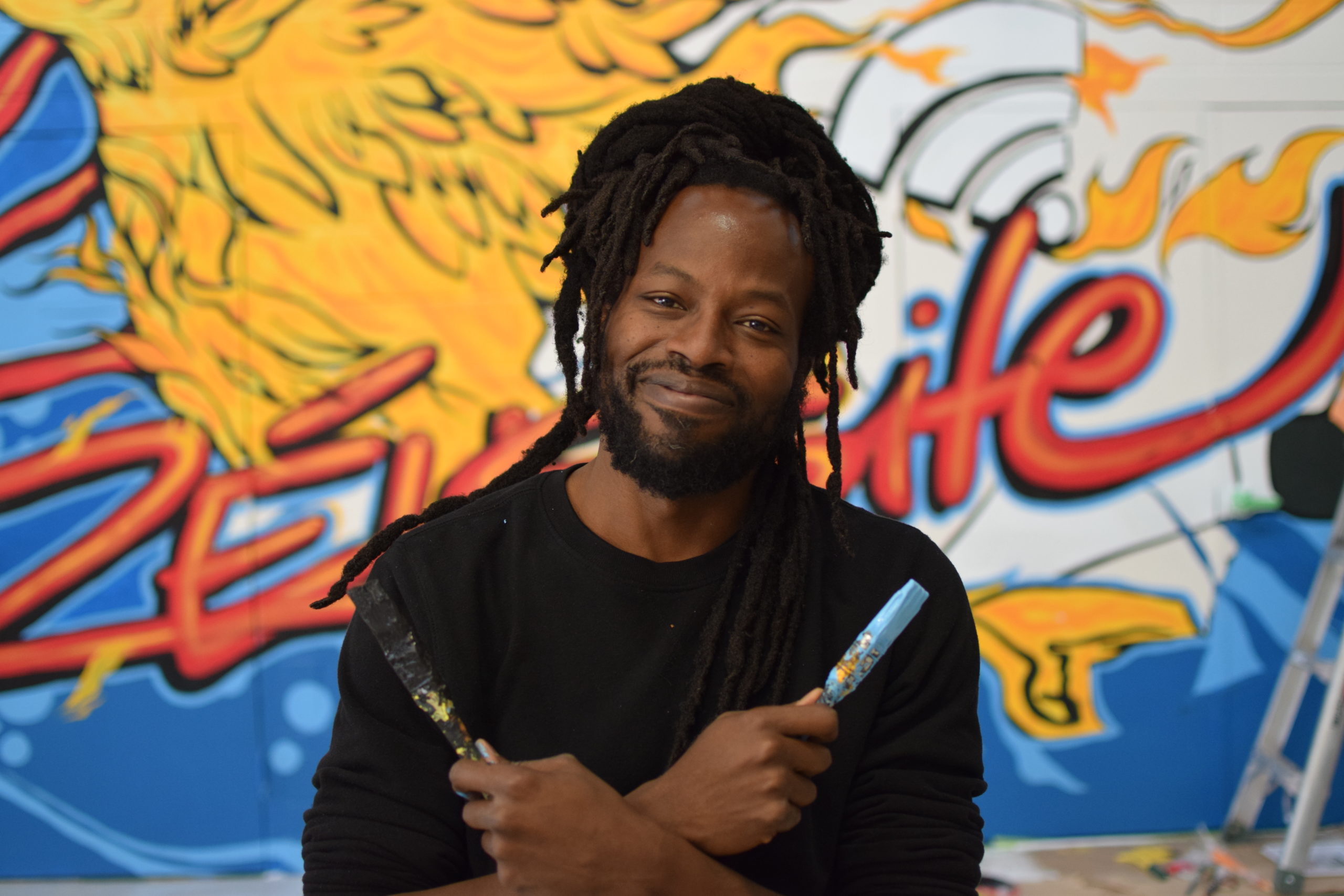 A Black man smiles in front of a colourful mural, holding two paintbrushes in an X across his chest. / Un homme Noir sourit devant une murale colorée en entrecroisant deux pinceaux devant son torse.