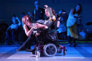 Two women dance, one in a wheelchair, while others dance in the background under blue tinted lights. / Deux femmes dansent, l’une d’entre elles en chaise roulante, pendant que d’autres dansent en arrière-plan sous une lumière bleue.