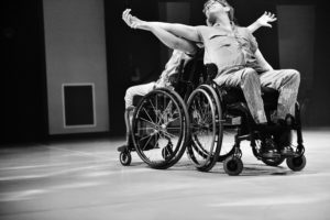 Two people in wheelchairs dance back to back in black and white, intertwining their arms. / Deux personnes en chaises roulantes dansent dos à dos en noir et blanc, en s’entrecroisant les bras.