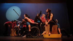 Performers in red, black, and blue dance together while others sit on set pieces or play instruments. / Des interprètes en rouge, noir et bleu dansent ensemble alors que d’autres d’entre eux sont assis sur des éléments du décors ou jouent des instruments.