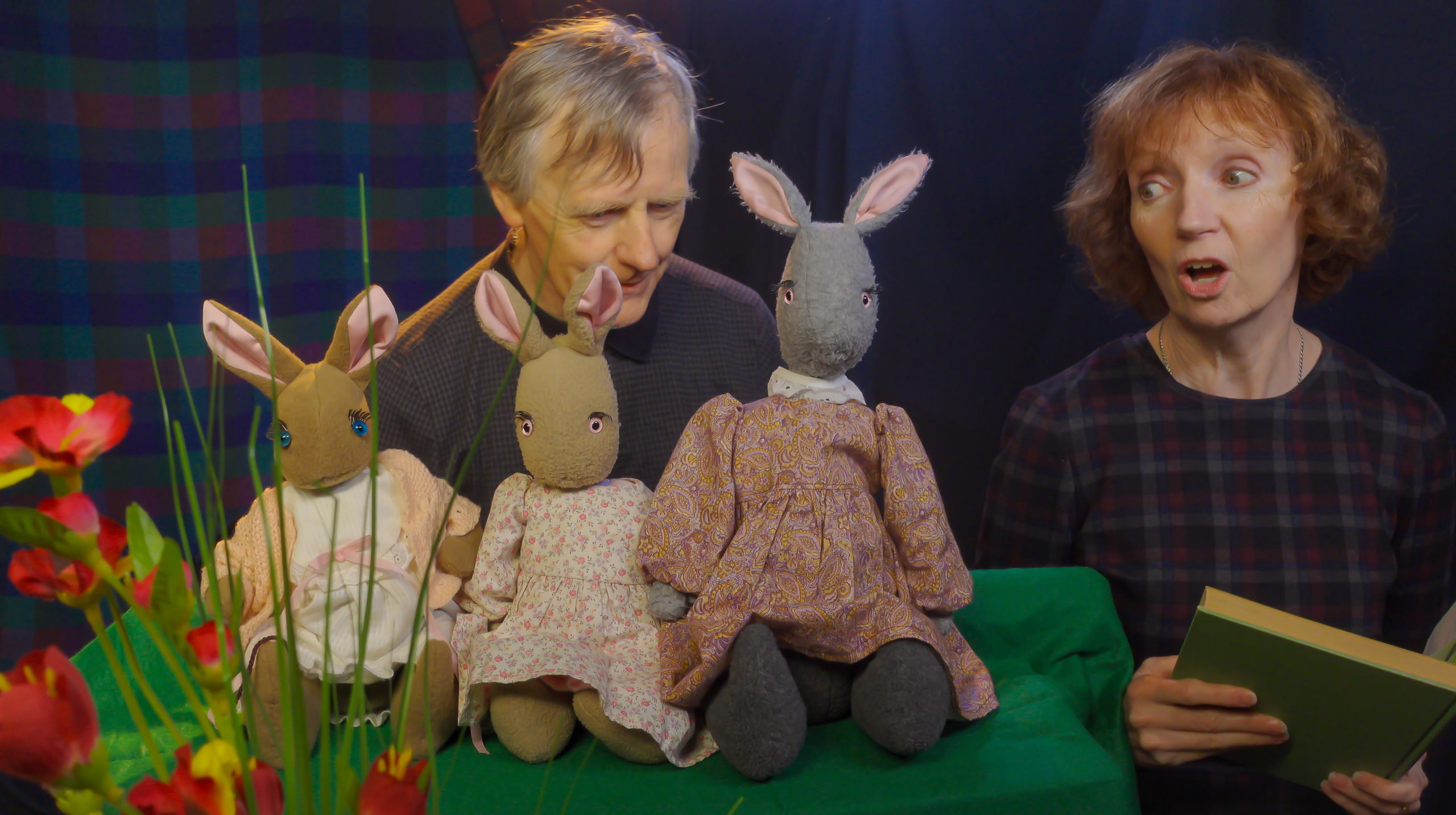 A White man and woman act, three rabbit puppets in dresses in front of them / Un homme et une femme Blanche jouent, trois marionettes de lapins en robes devant eux.