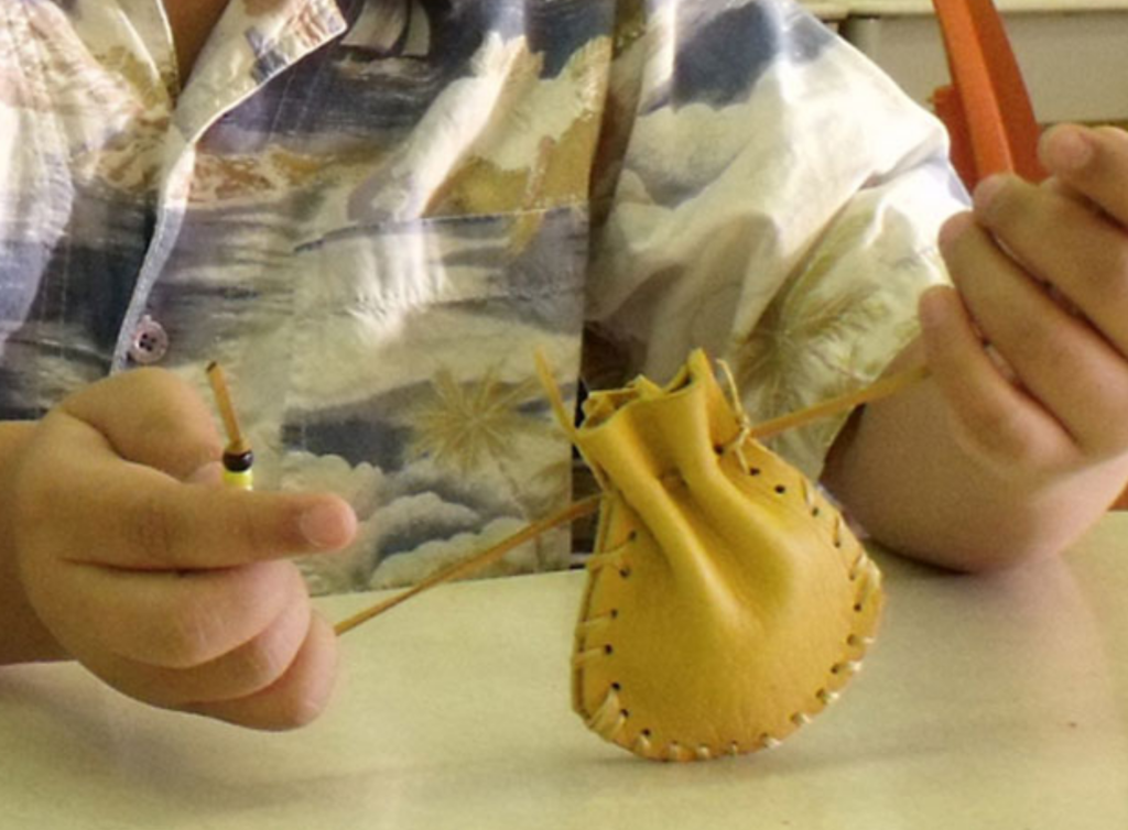 A close up of child’s hands pulling the drawstrings of a light brown leather medicine pouch to close it. / Un gros plan des mains d’un enfant qui tirent les cordons d’un sac de plantes médicinales, fait de cuir marron pâle, pour le refermer.